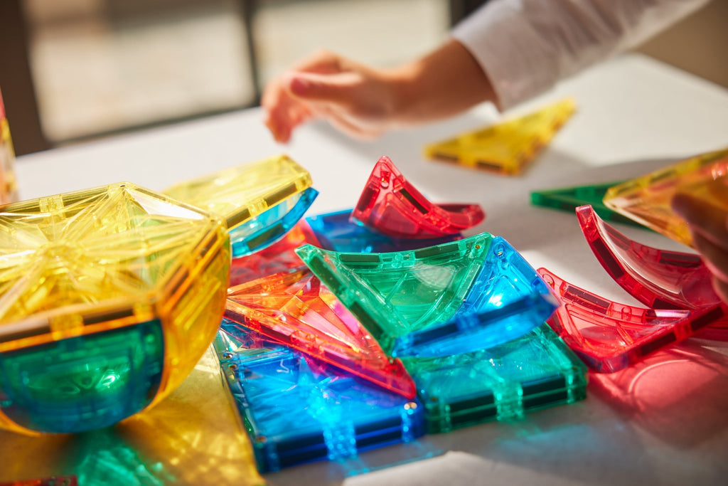 Bambino che gioca con costruzioni magnetiche traslucide Cleverclixx colorate su un tavolo alla luce del sole.