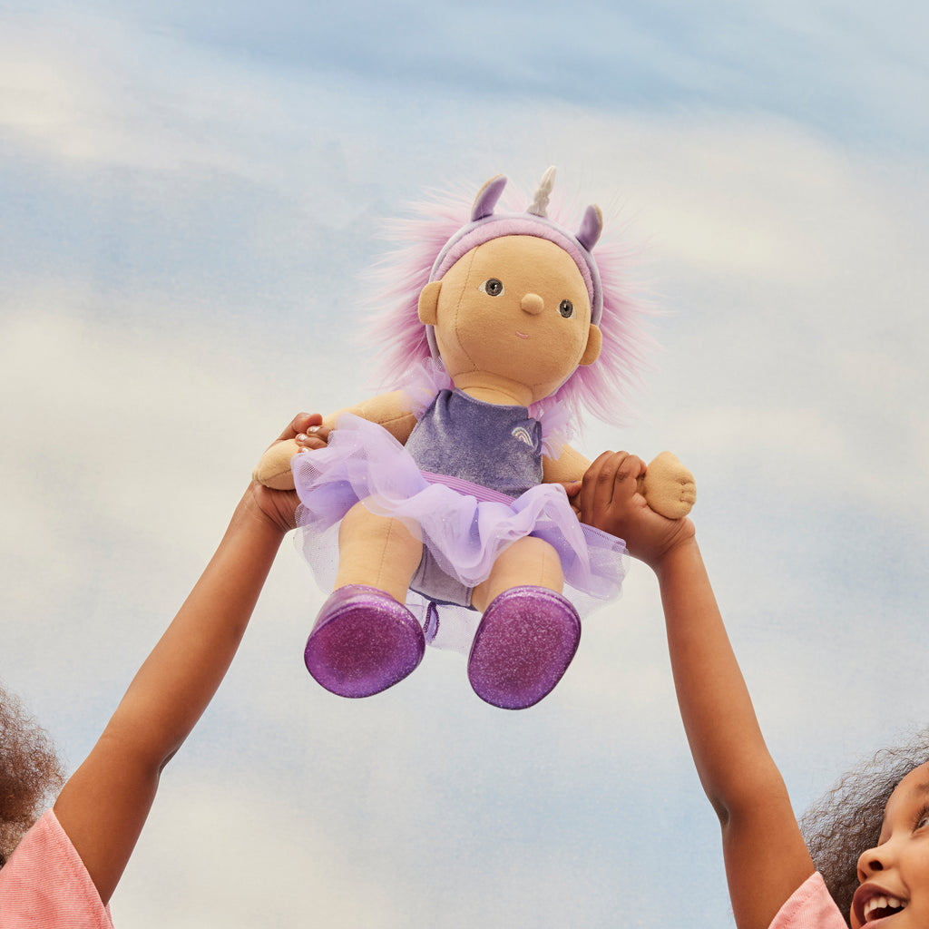 Bambina solleva in alto la bambola Dream Dinkum Violet di Olli Ella contro il cielo azzurro
