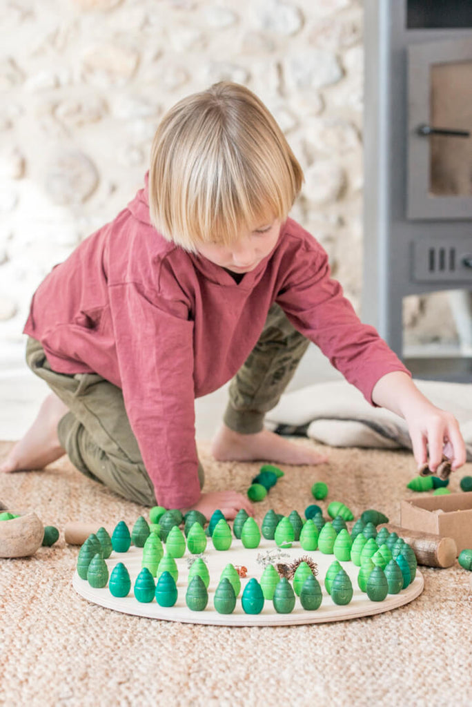 Bambino che gioca con il set Grapat Mandala Tree creando un mandala verde su tappeto naturale, vista dall’alto