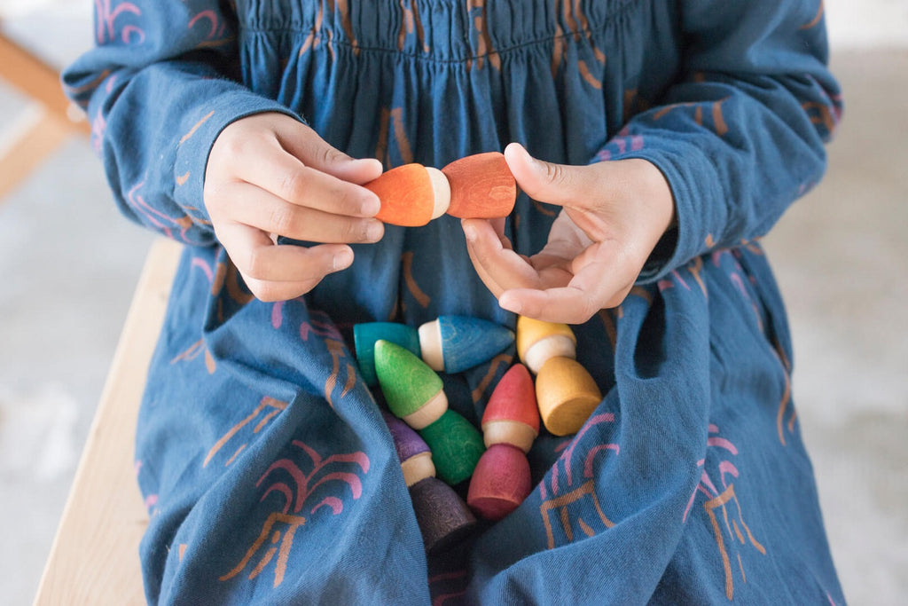 Bambina che gioca tenendo due figure in legno Grapat Rainbow Tomten, con altri Tomten appoggiati sul vestito