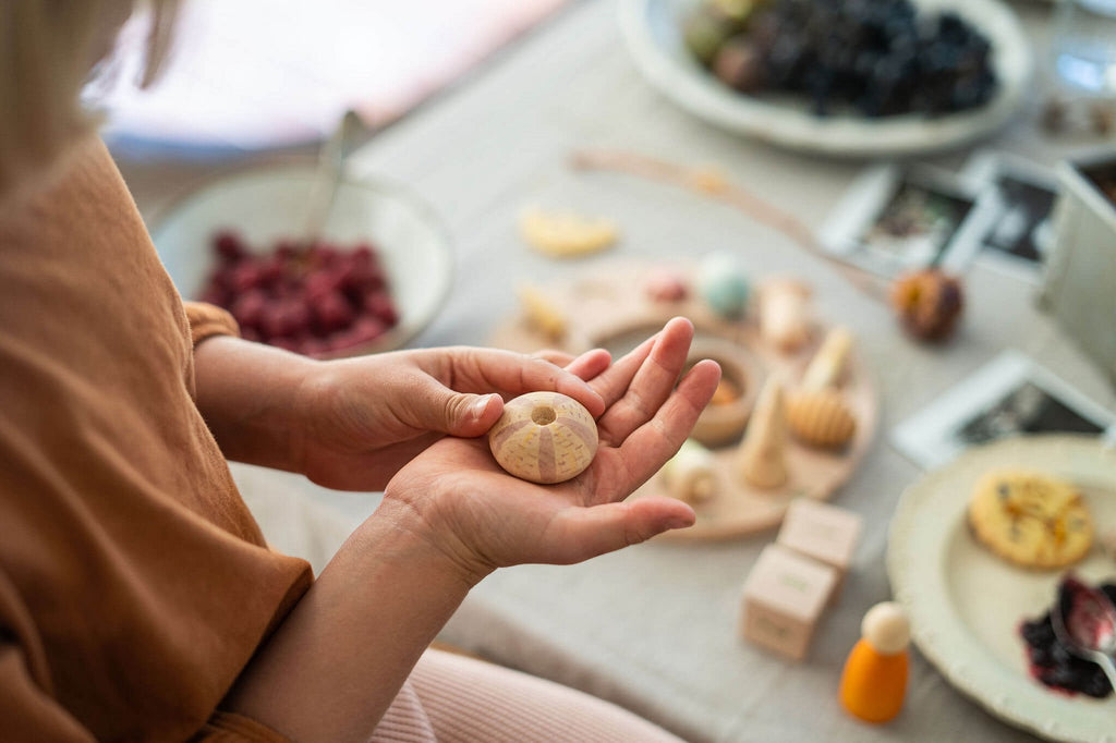 Bambina che osserva un elemento in legno del set Grapat Wonders, seduta al tavolo con altri oggetti sensoriali naturali.